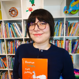 A person wearing glasses and a dark top holds an orange book titled “Huevos verdes con jamón” in front of shelves filled with children’s books and colorful decorations.