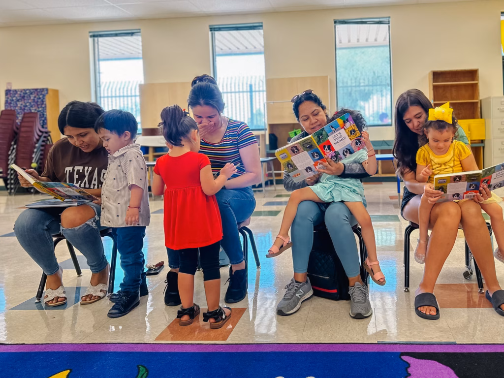 Four adults and four young children sit in a brightly lit room, looking at colorful picture books; the visible book covers read “LET’S FIND MOM!”