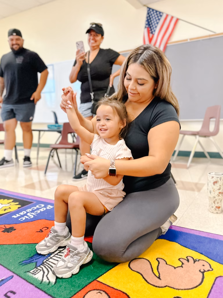 An adult woman kneels on a colorful classroom rug while holding a smiling young child who raises one hand. Two adults stand in the background, one holding a phone. An American flag, chairs, tables, and a blank board are visible in the room.