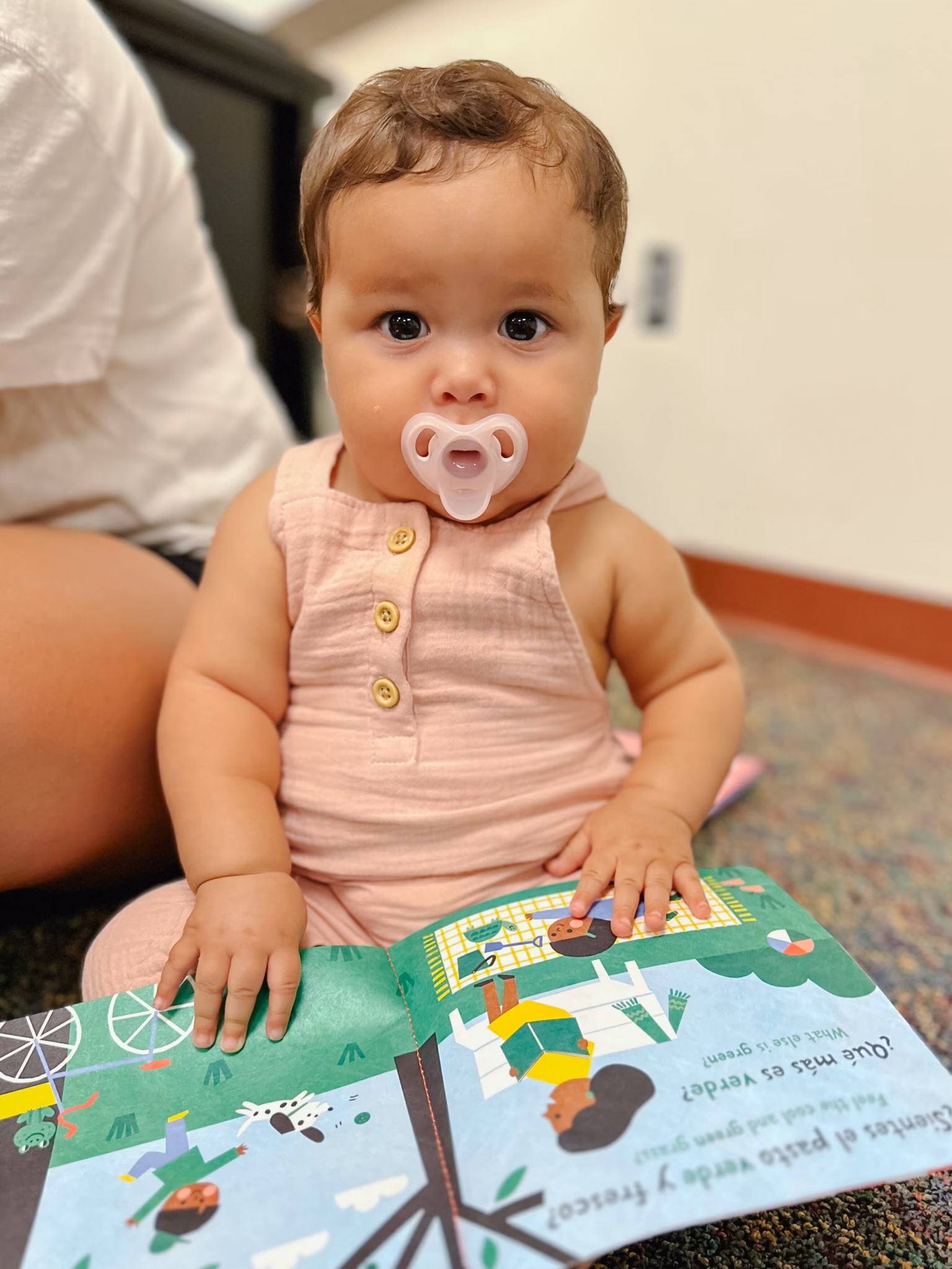 Baby with brown hair and a translucent pacifier, wearing a pale pink romper and seated while holding an open illustrated book. Part of an adult is visible beside the baby.