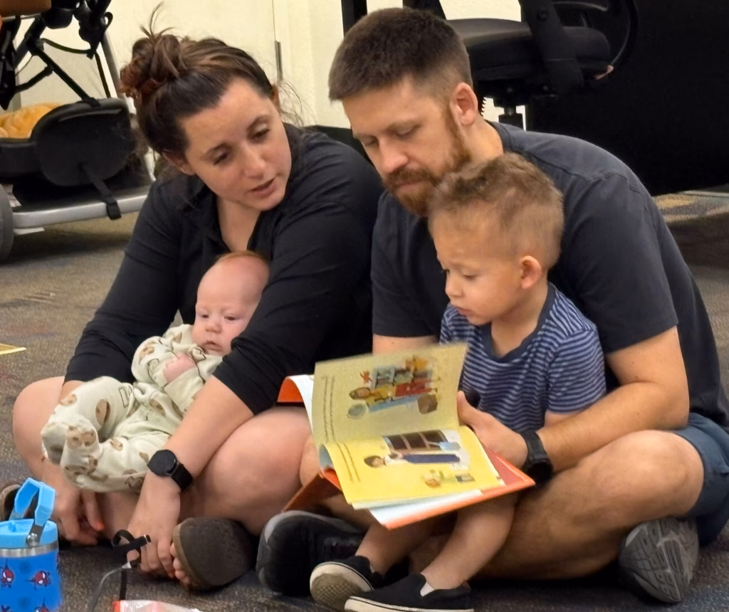 Two adults sit on the floor with two young children; one adult holds a baby while the other helps a toddler look at a picture book. Chairs and bags are visible in the background.