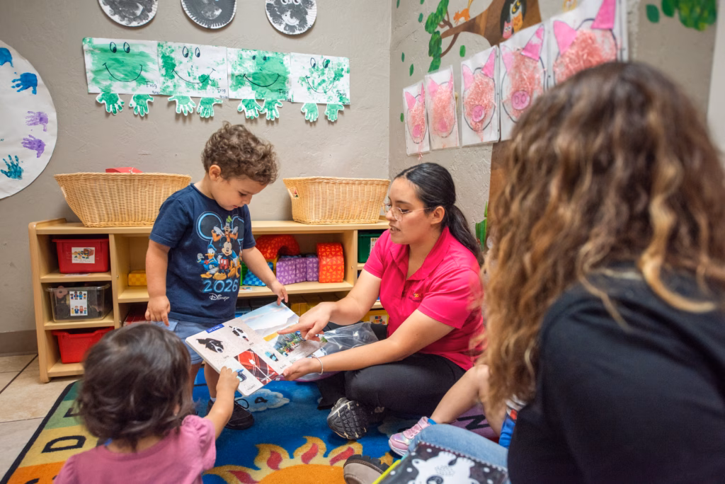 An adult in a pink shirt sits on a classroom rug, showing an illustrated book to young children. Colorful storage shelves and frog and pig artwork decorate the room.