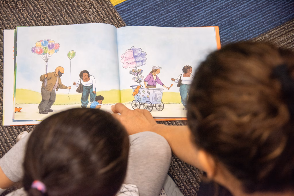 Two children look at an open illustrated picture book showing people outdoors, including a balloon seller and a colorful treat cart.