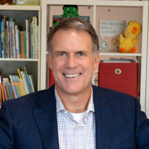 Smiling man in a dark blue blazer and checkered shirt, photographed in front of shelves filled with books, toys, red storage bins, and a sign reading “The Duckling Gets a Cookie!?”